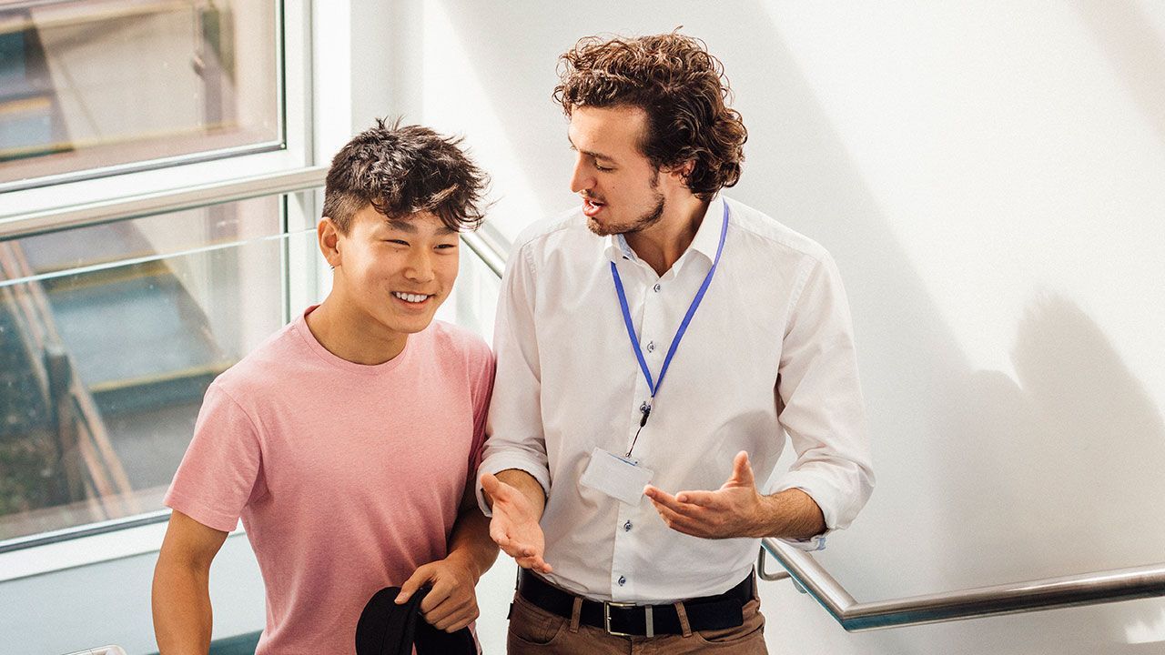 A young man and a teacher, both smiling, walk down stairs in a school.