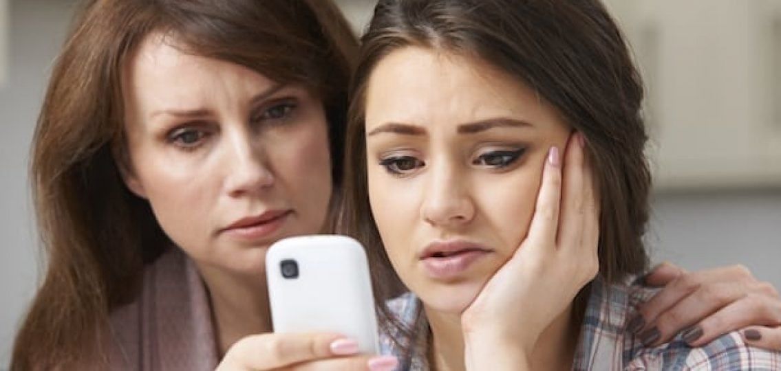 Mother comforting daughter looking at a phone, both with concerned expressions.