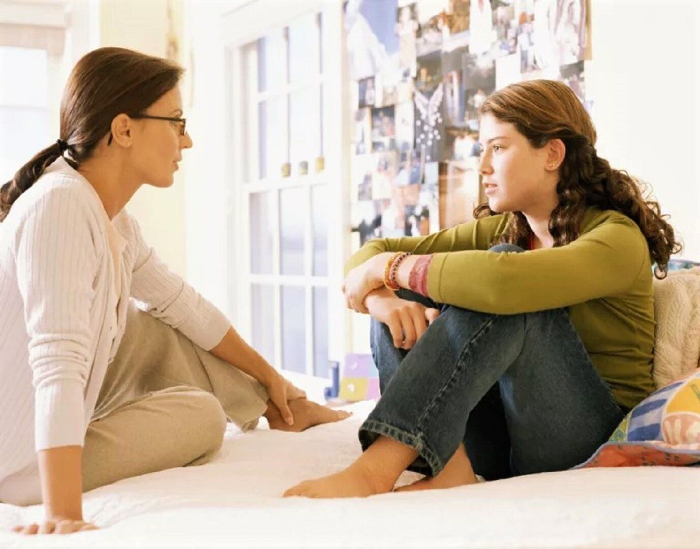Woman in glasses talking to a teen in a bedroom. Both are sitting on a bed, engaged in conversation.