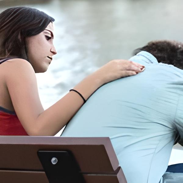 Woman comforts a slumped man on a park bench; she places a hand on his shoulder.