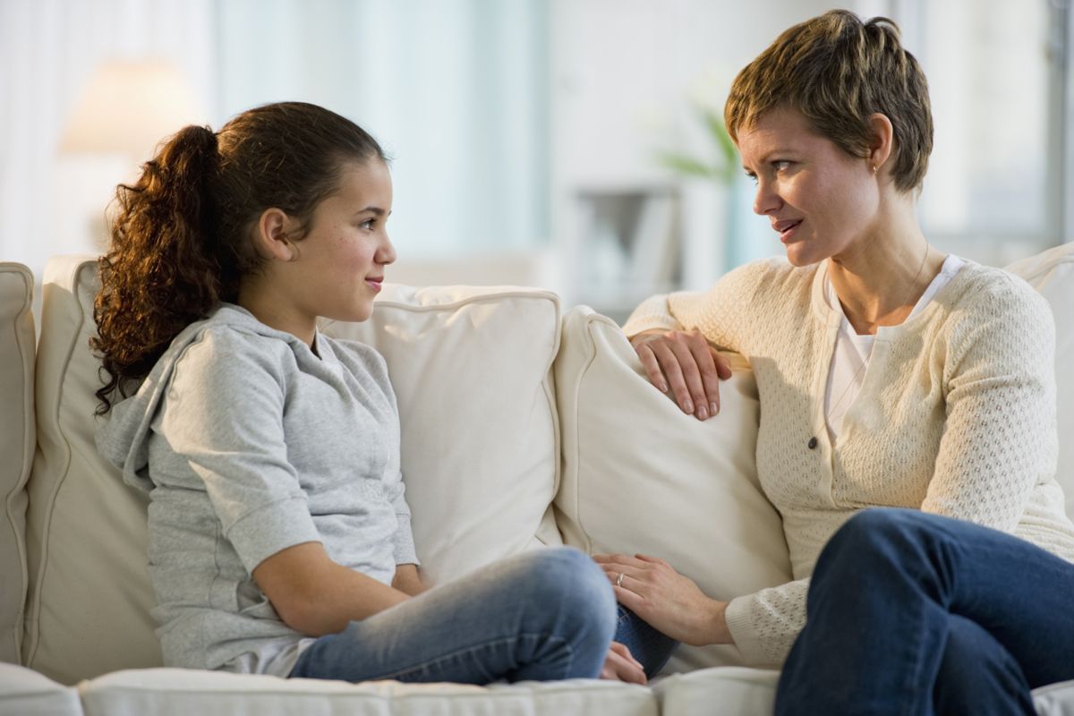Mother and daughter sit on a couch, talking. The woman has her hand on the girl’s knee, both look thoughtful.