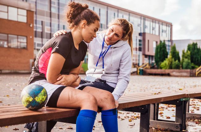 A field hockey player is comforted by a coach on a bench after an injury.