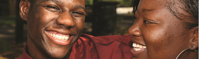 Two people of color smiling, laughing. Man in red shirt, woman next to him. Outdoor setting, warm tones.