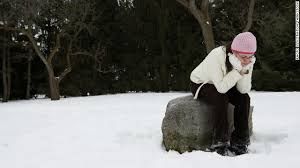 Woman in winter clothes sits on a snowy rock, looking down with a sad expression.