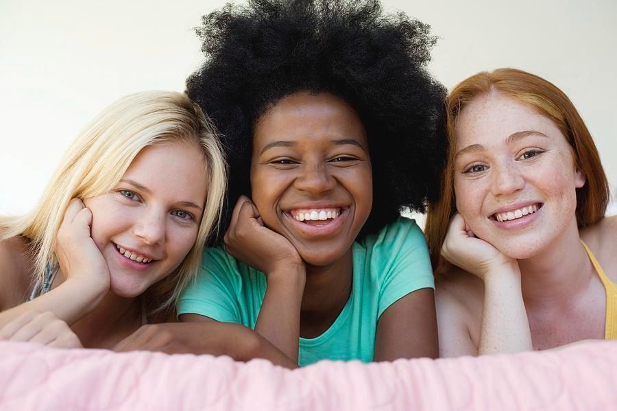 Three diverse young women smiling, resting chins on hands, lying on a pink blanket.