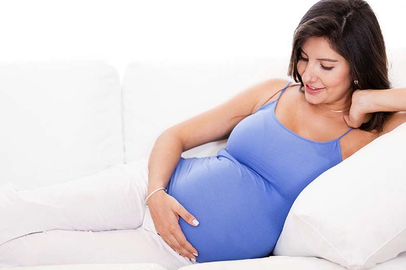 Pregnant woman in blue tank top, smiling, rests on white couch, hand on belly.