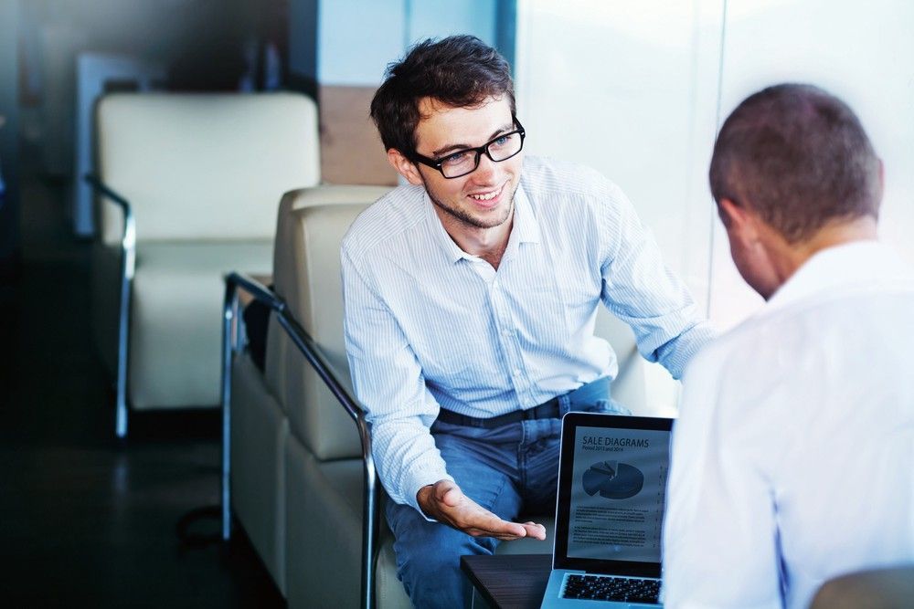 Two men in office setting, one showing laptop with charts, the other facing him. One is pointing and smiling.