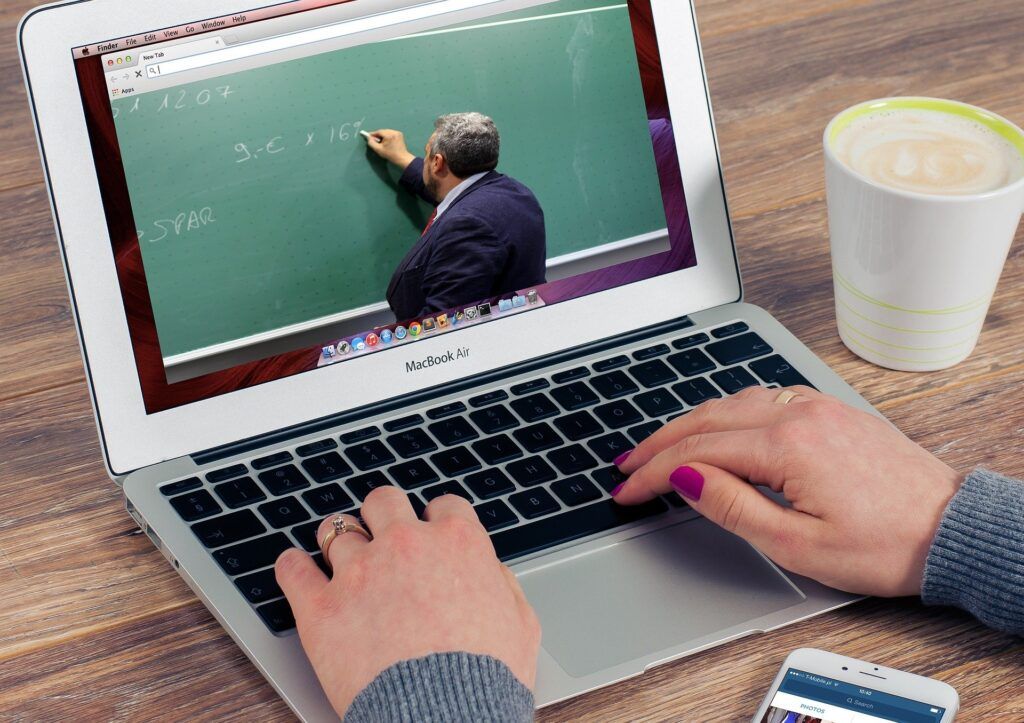 Person using laptop for online class, watching a teacher write on a chalkboard. Coffee and phone are nearby.