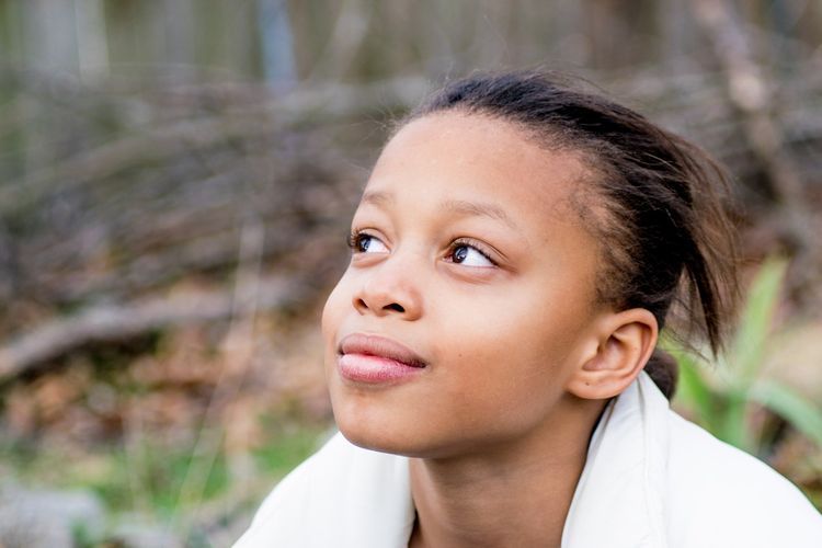 Girl looking up with a thoughtful expression, light skin, outdoors with trees blurred in the background.