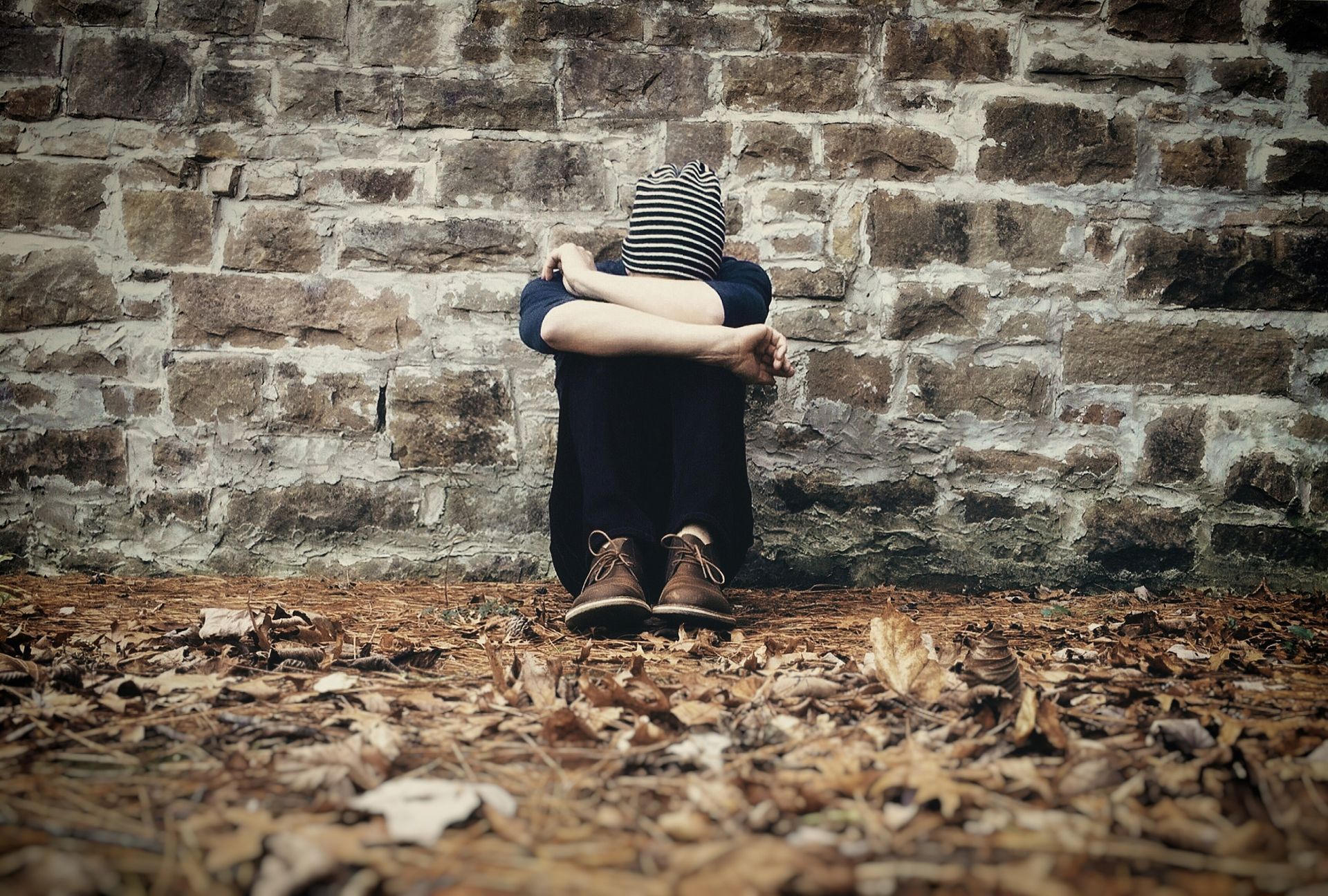 Person in striped hat sits against brick wall, arms wrapped around knees.