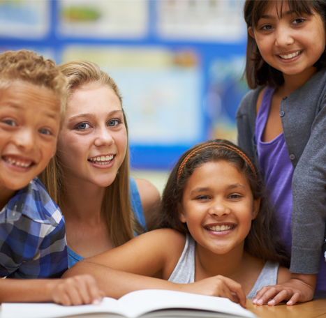 Group of smiling children gathered around an open book in a classroom.