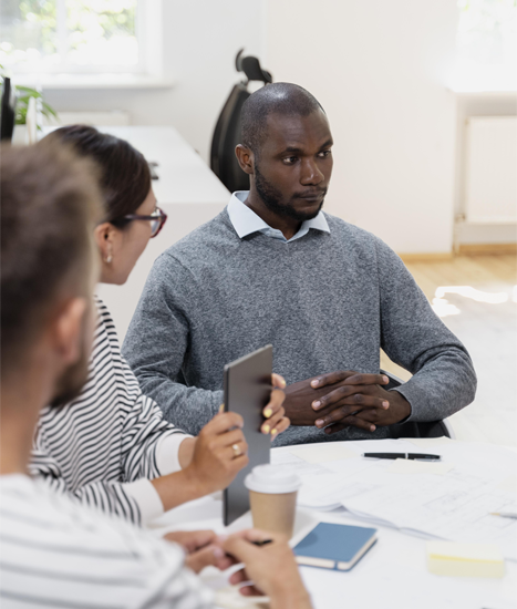 Man in gray sweater at a meeting, looking intently. Others present at a white table.