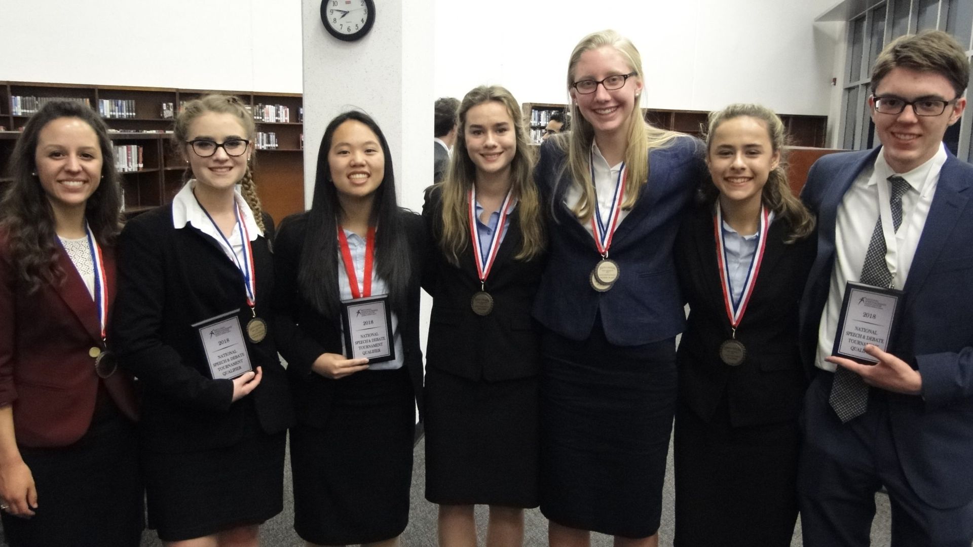 Group of students in suits holding awards, smiling for a photo in a library.
