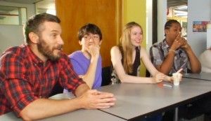 Four people seated at a table indoors, smiling, listening to a person with a beard speak.