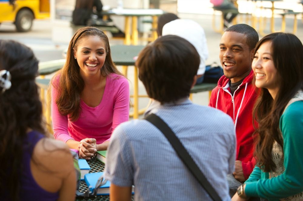 Group of diverse students laughing and conversing outdoors at a table.