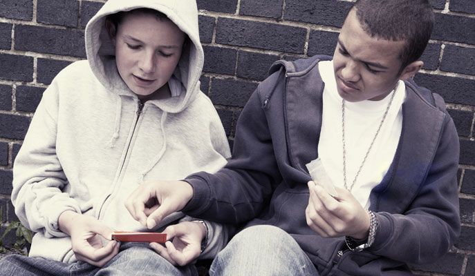 Two young males, one white and one Black, are sitting together; they appear to be rolling something between their fingers near a brick wall.