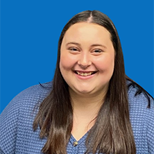 Woman with long dark hair smiles at the camera against a blue background, wearing a blue patterned top.