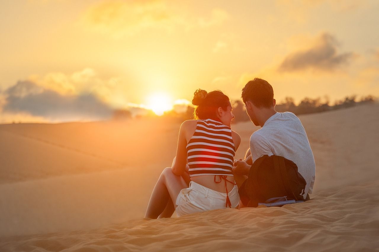Couple sitting on a sand dune, watching the sunset. Golden light, silhouetted figures.