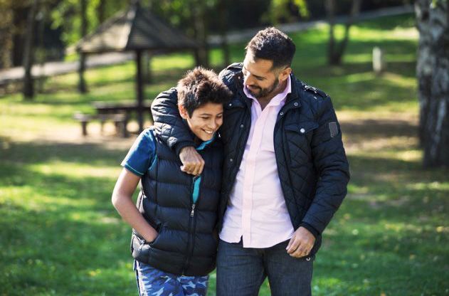 Father and son walking arm-in-arm in a park; the father smiles, the son looks down smiling.