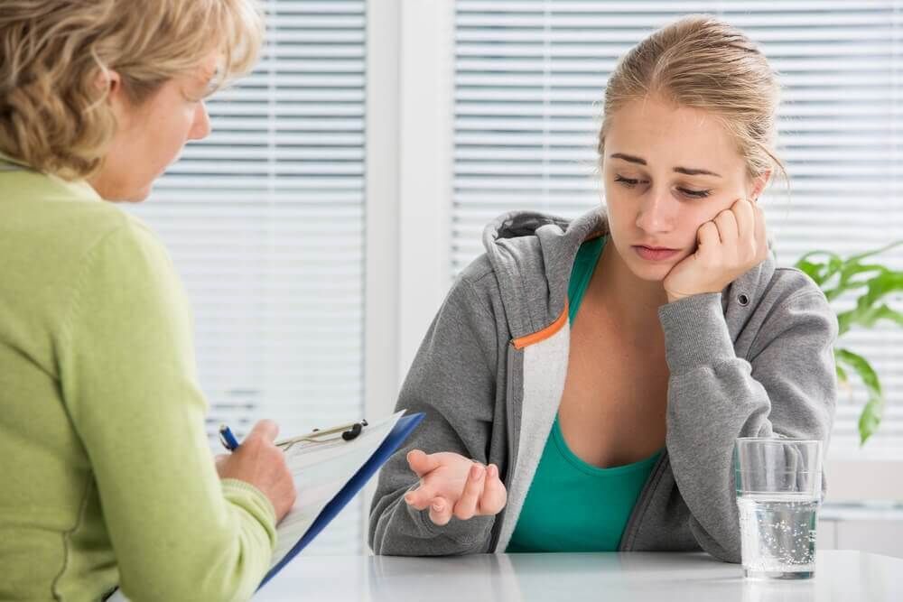 Young woman talking to a therapist, looking sad, in a bright office setting.