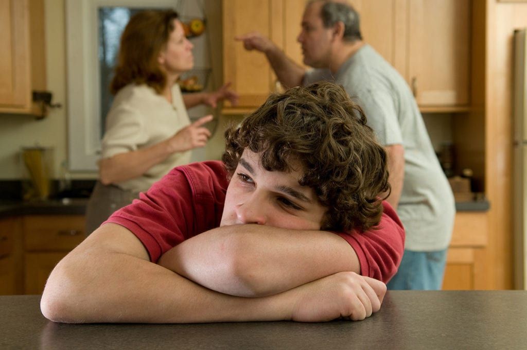 Teenage boy with arms on a table, parents arguing in the background in a kitchen.