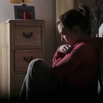 A young person sits in a dimly lit room, looking down, arms wrapped, appearing sad near a nightstand.