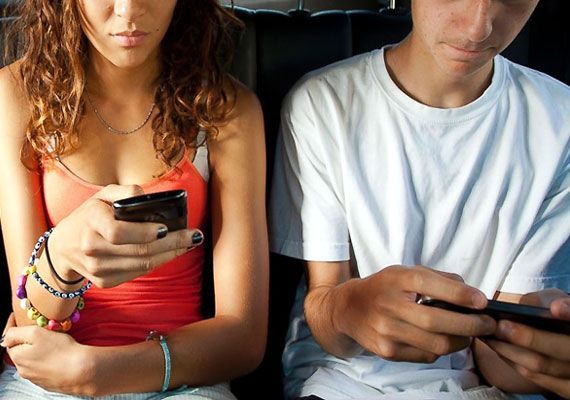 Girl and boy using phones, inside a car. Girl wears orange top, boy white shirt. Both have focused expressions.