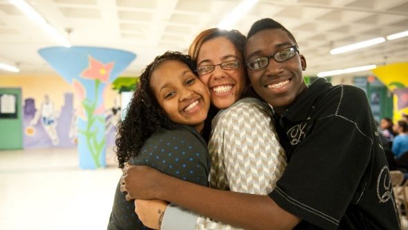 Three people, smiling and hugging. A woman and two young adults in a brightly colored school hallway.