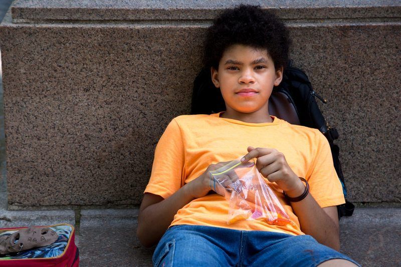 Boy with curly hair in orange shirt, sitting against a stone wall, holding a plastic bag with food.