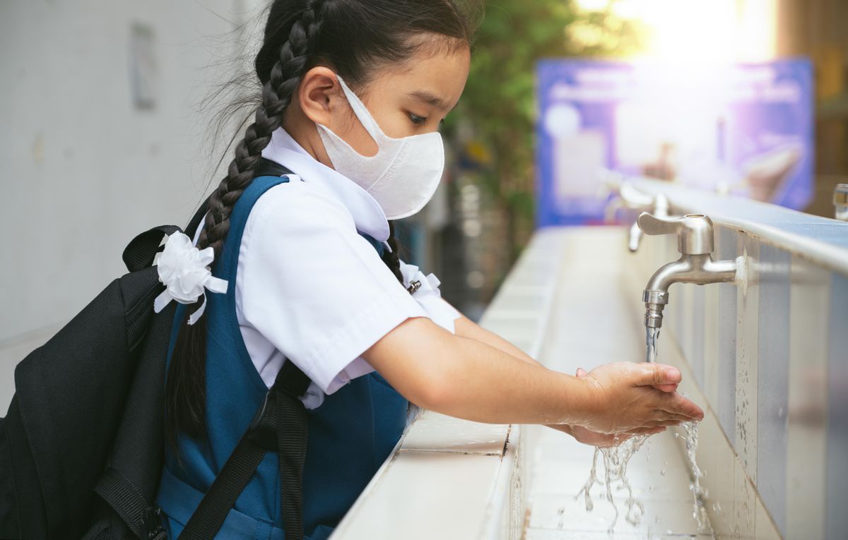 Girl in school uniform and mask washes hands at an outdoor sink.