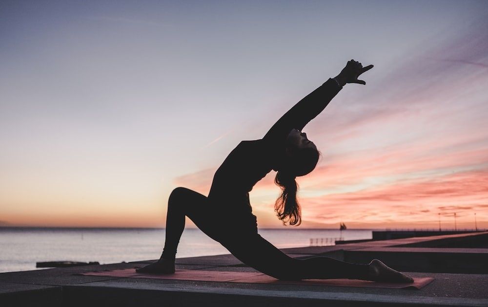 Silhouette of a person doing yoga outdoors at sunset, one arm raised, the ocean in the background.
