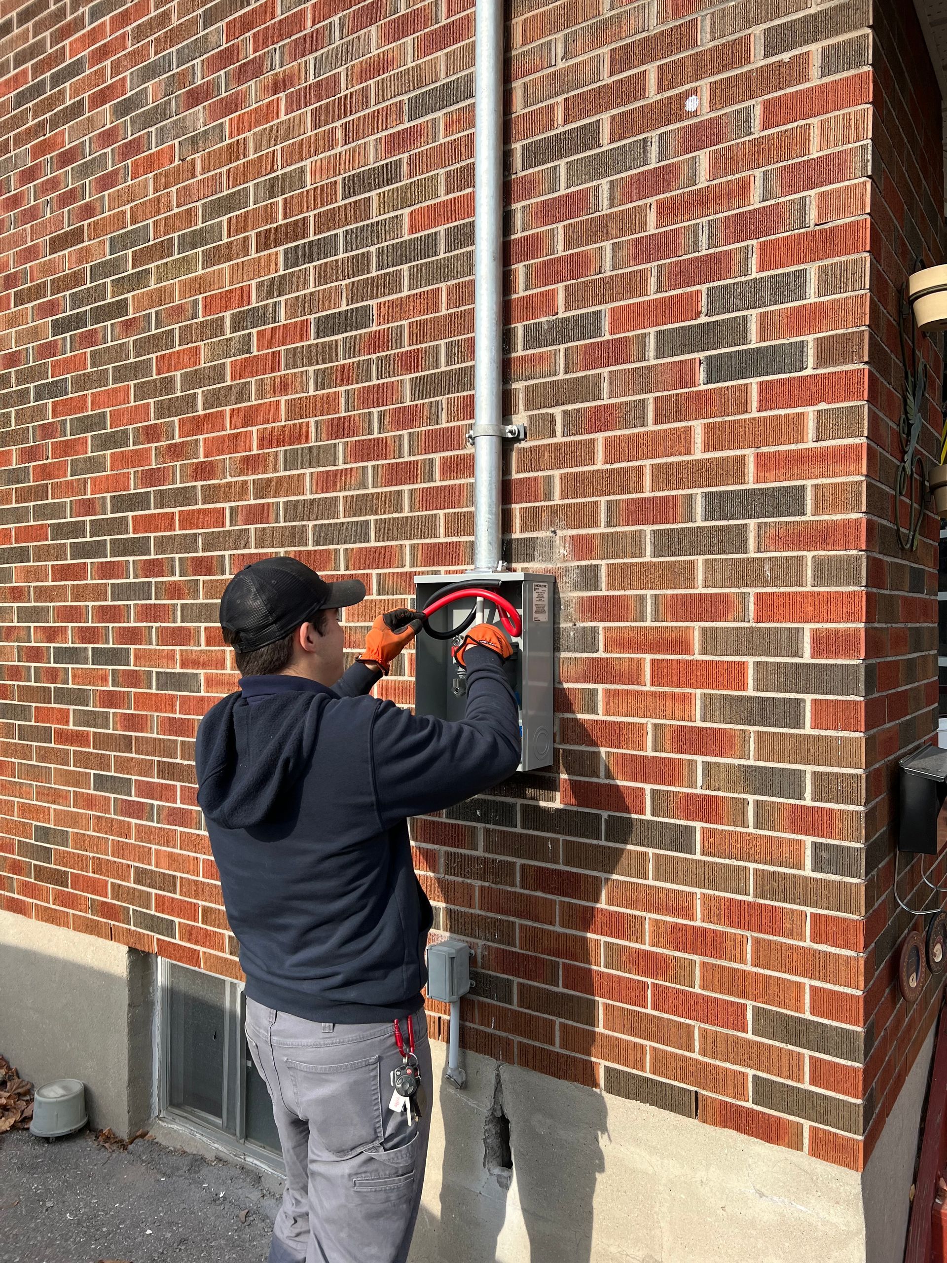 Electrician working on electrical box, attached to brick wall. He wears a cap and orange gloves.