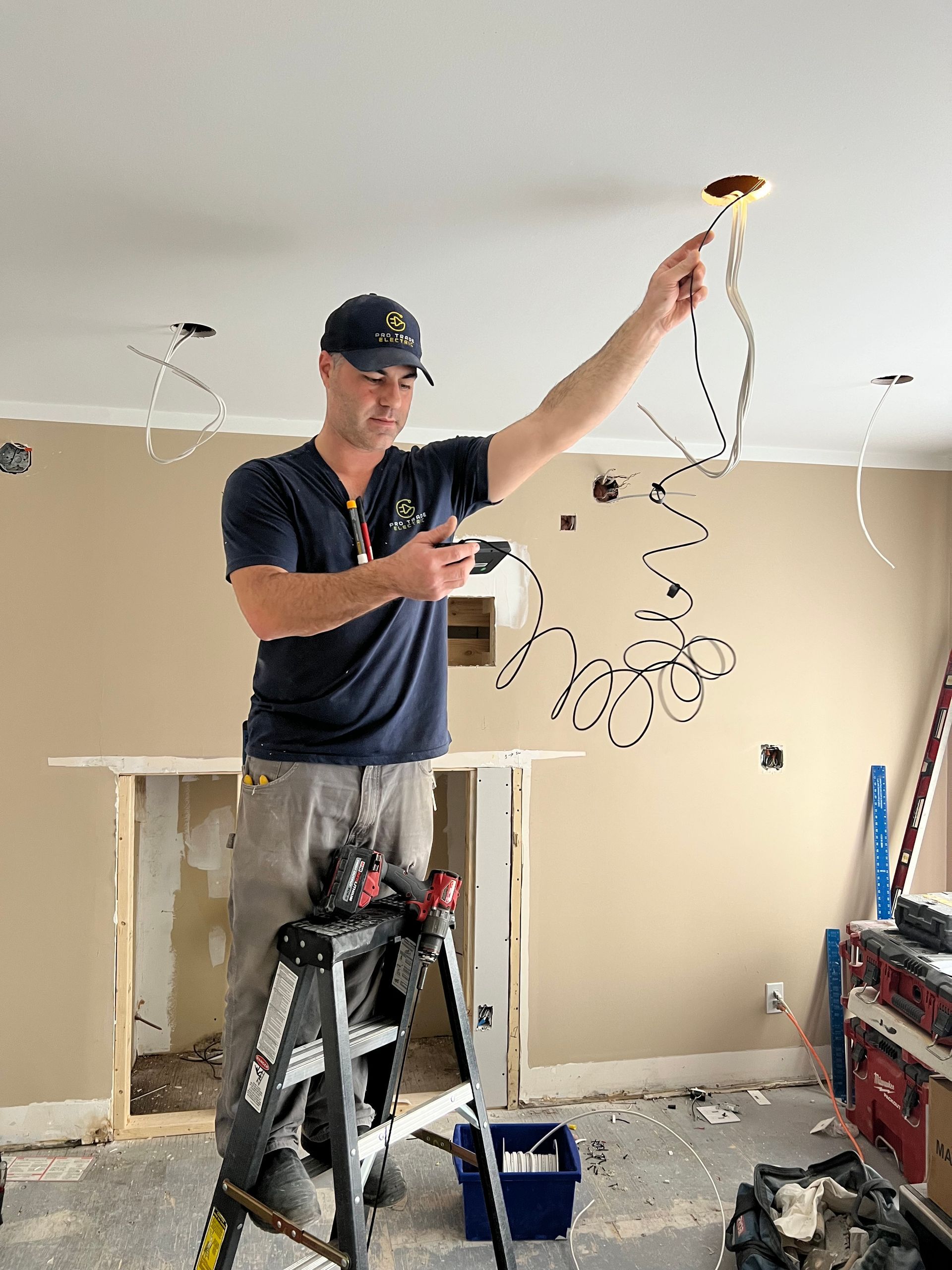 Man on ladder, holding wires, inspecting electrical work in a room under construction.