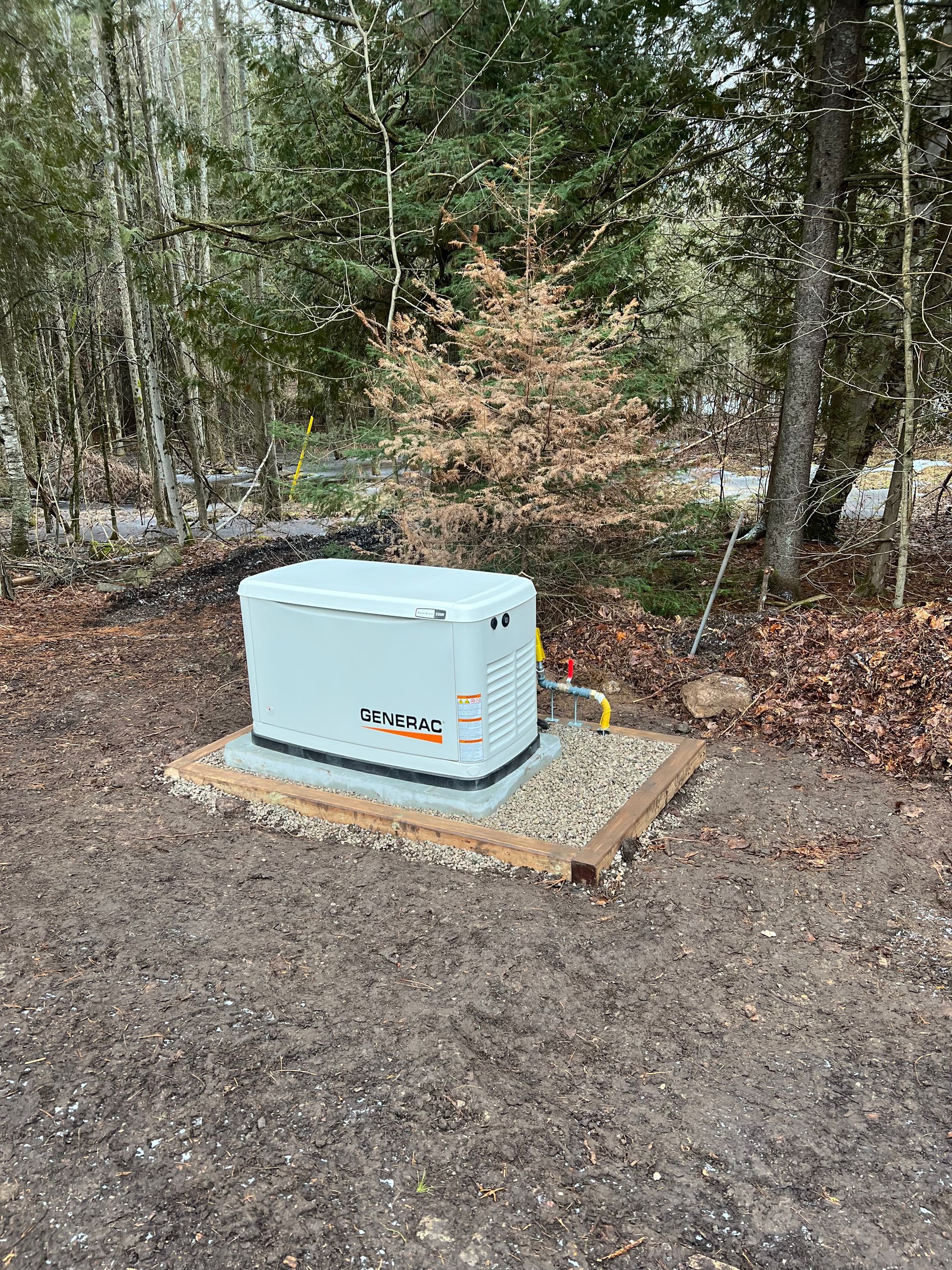 Generator on a gravel pad near trees. The generator has a yellow fuel line and a concrete border.