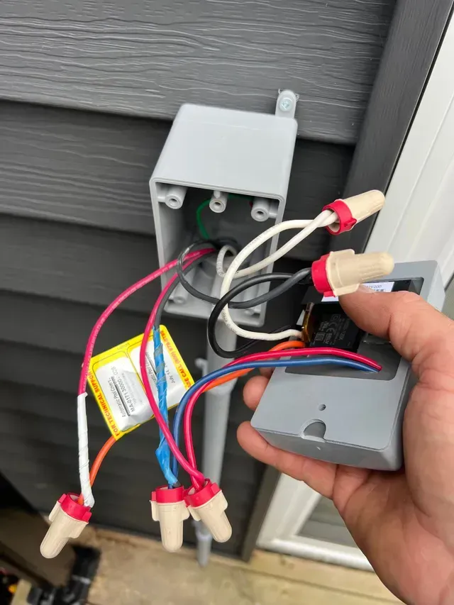 Hand holding electrical wiring box. Gray box and wires with red wire connectors. Outdoors, siding visible.