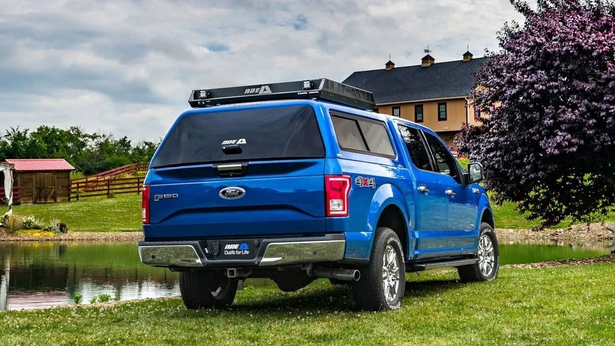 Blue pickup truck parked on grass beside a pond with a house in the background