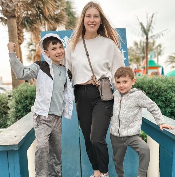 Woman and two boys posing by a blue structure. Woman in white sweater, black pants, boys in hoodies, palms in background.
