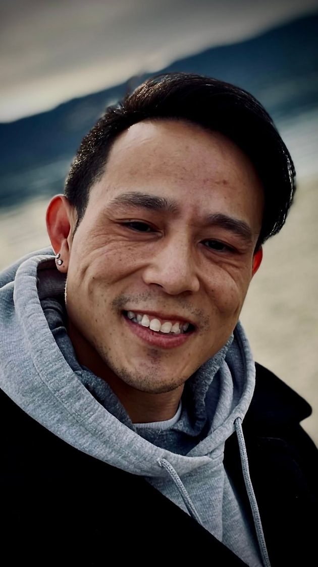 Man smiling, wearing a gray hoodie and black coat, outdoors with blurred background.