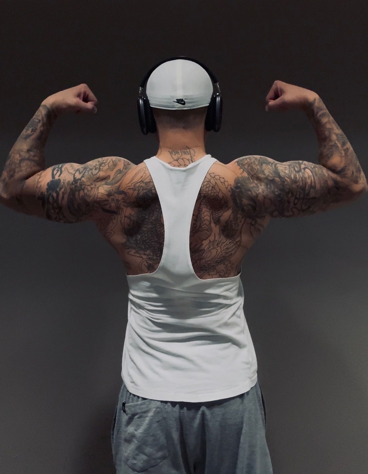 Man flexing biceps, wearing a white tank top, tattoos on arms, and grey sweats, headphones, against a grey background.