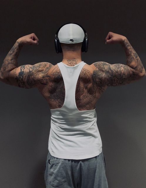 Man flexing biceps, wearing a white tank top and gray shorts. Tattooed arms and back. Gray background.