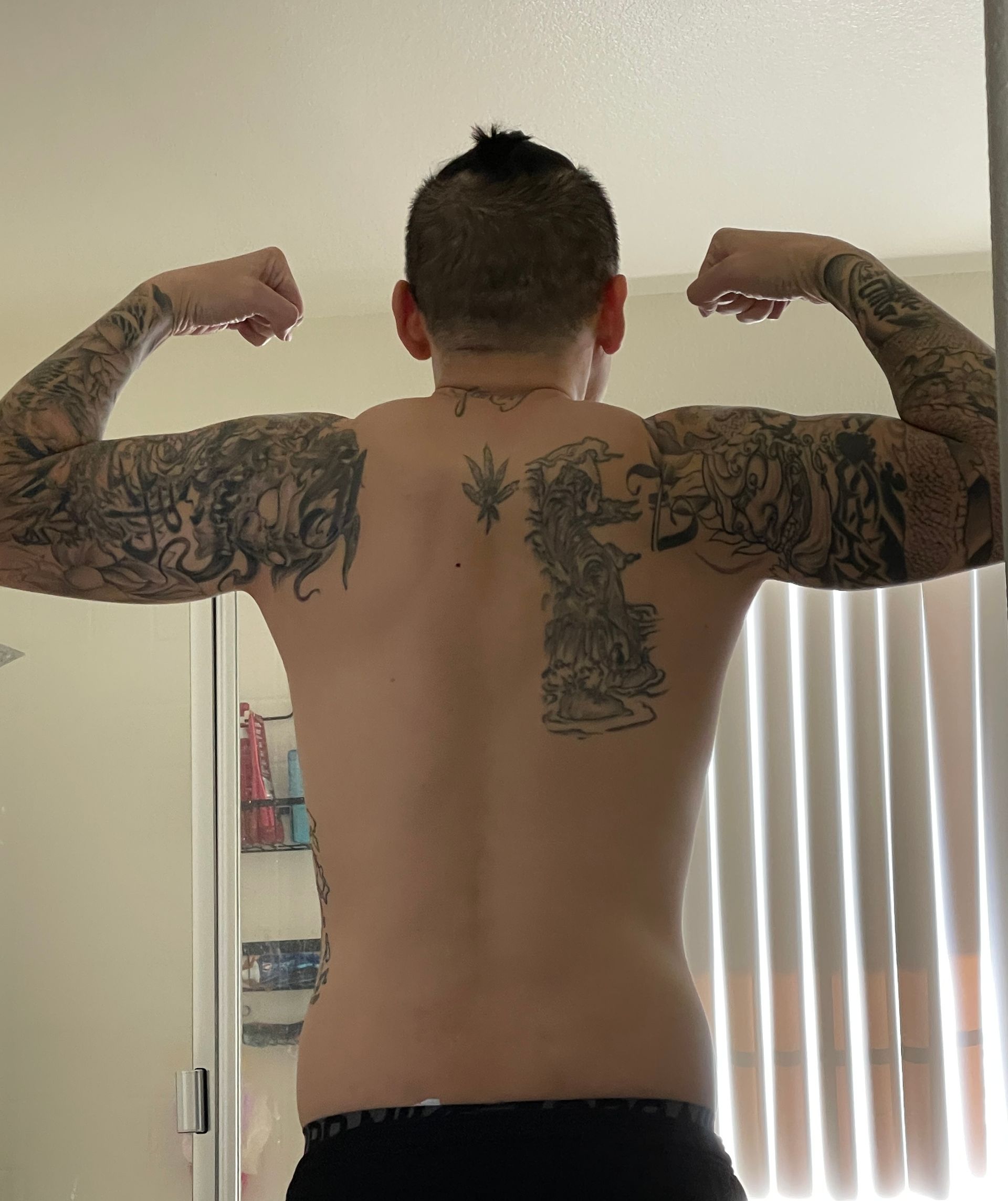 Person flexes biceps, showing arm and back tattoos. Short hair, indoors, flexing towards viewer.