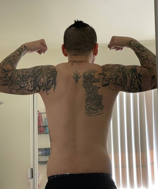 Person flexing arm muscles, displaying back tattoos, standing indoors near a window.