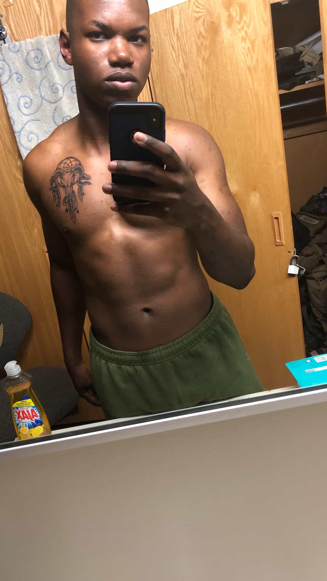 Person taking a mirror selfie, shirtless, tattoo on chest, green sweatpants. Bathroom setting.