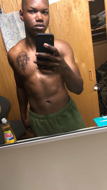 A person shirtless, with a tattoo on their chest, holding a phone in front of a bathroom mirror.