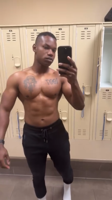 Man with muscular torso takes a gym selfie in front of lockers, wearing black pants and white socks.