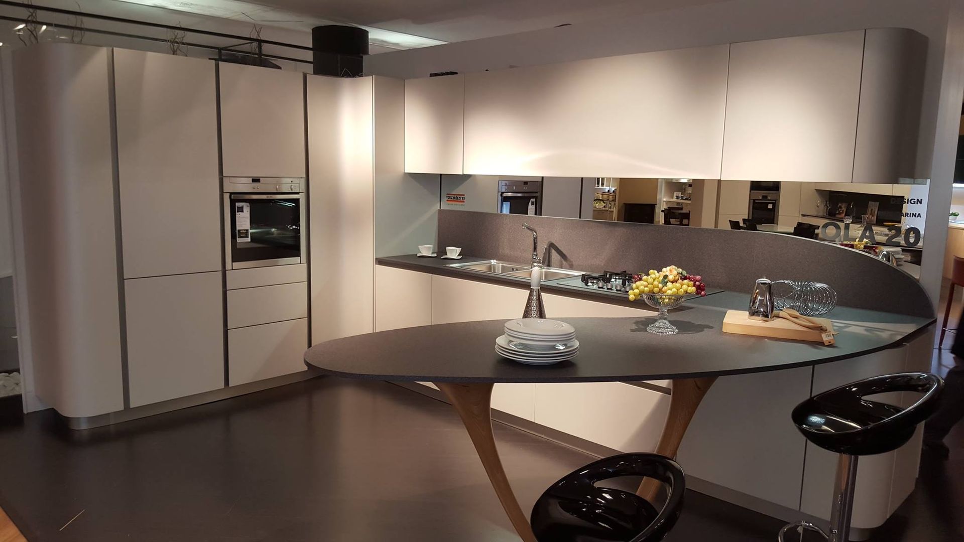 Cucina moderna con isola con piano di lavoro curvo, mobili bianchi, isola grigio scuro e sgabelli da bar neri.