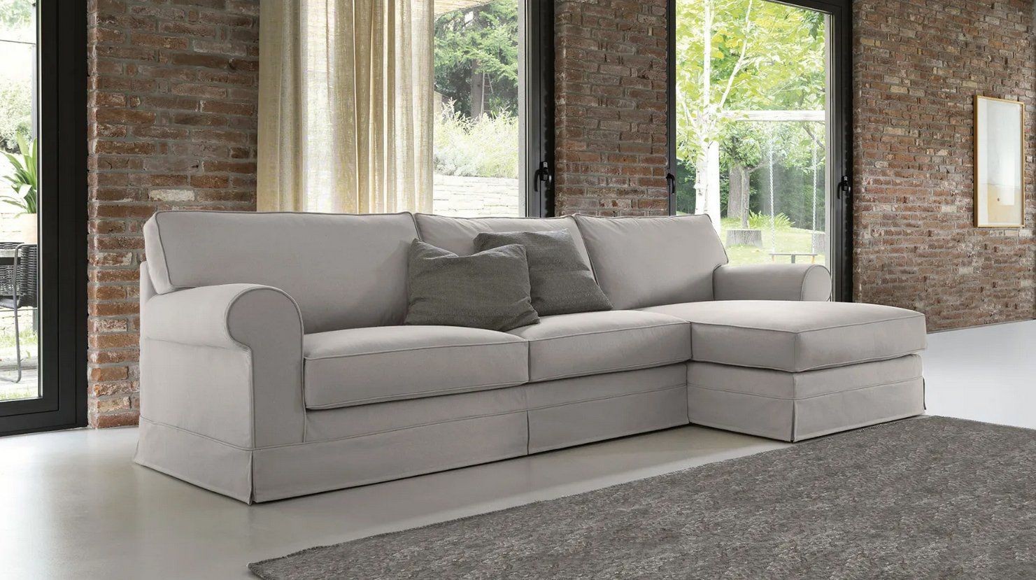 Divano componibile beige in una stanza con una parete in mattoni, ampie finestre e un tappeto grigio.