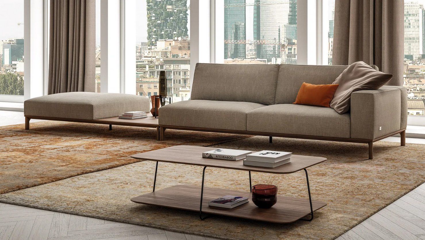 Soggiorno con divano color marrone chiaro, pouf e tavolino da caffè su un tappeto a pelo lungo, ampie finestre.
