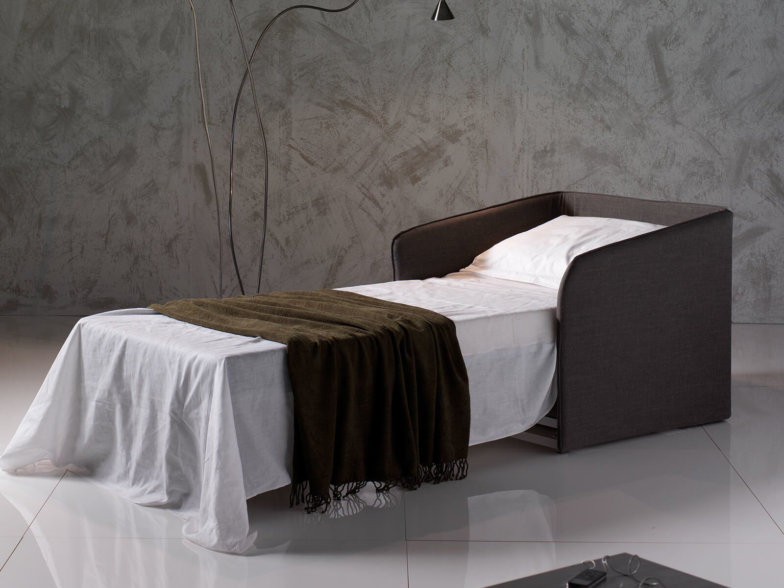Divano letto marrone con lenzuola bianche, coperta verde oliva e moderna lampada da terra.
