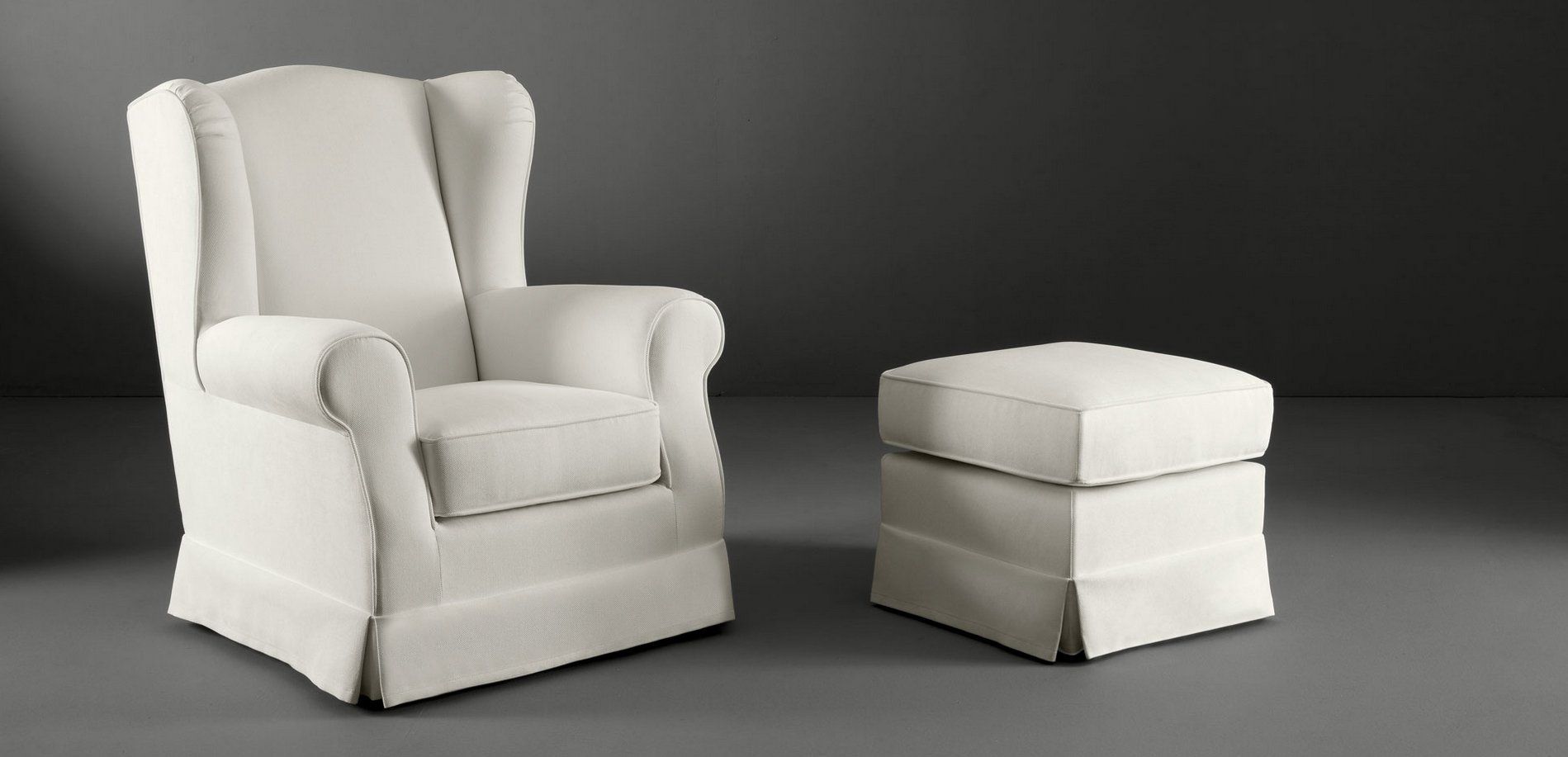 Poltrona con schienale alto color bianco sporco e pouf coordinato su sfondo grigio.